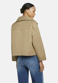 Vrouw met een knot in haar haar, gekleed in een beige cropped jasje en blauwe spijkerbroek, staand tegen een witte achtergrond en zijwaarts kijkend.