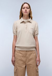 Beige lyhythihainen polo-paita, jossa on ribbitetty helma, kolme nappia ja kohokuvioitu logolla varustettu rintatasku. Yhdistetty vaaleanruskeisiin cargo-housuihin.