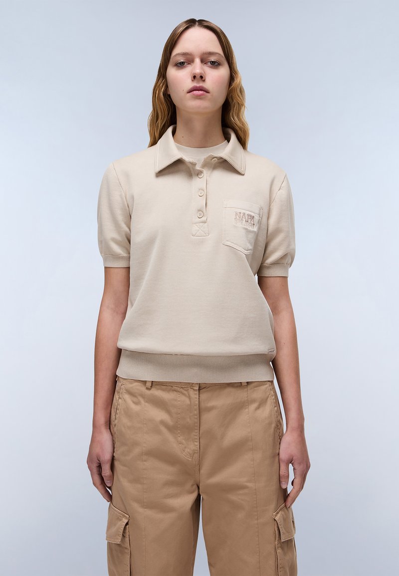 Beige lyhythihainen polo-paita, jossa on ribbitetty helma, kolme nappia ja kohokuvioitu logolla varustettu rintatasku. Yhdistetty vaaleanruskeisiin cargo-housuihin.
