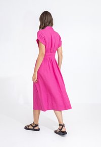 Růžové lněné šaty s krátkými rukávy, zavinovací pas a midi délka. Nosí se s černými plochými sandály, které ukazují jednoduchý a elegantní design.
