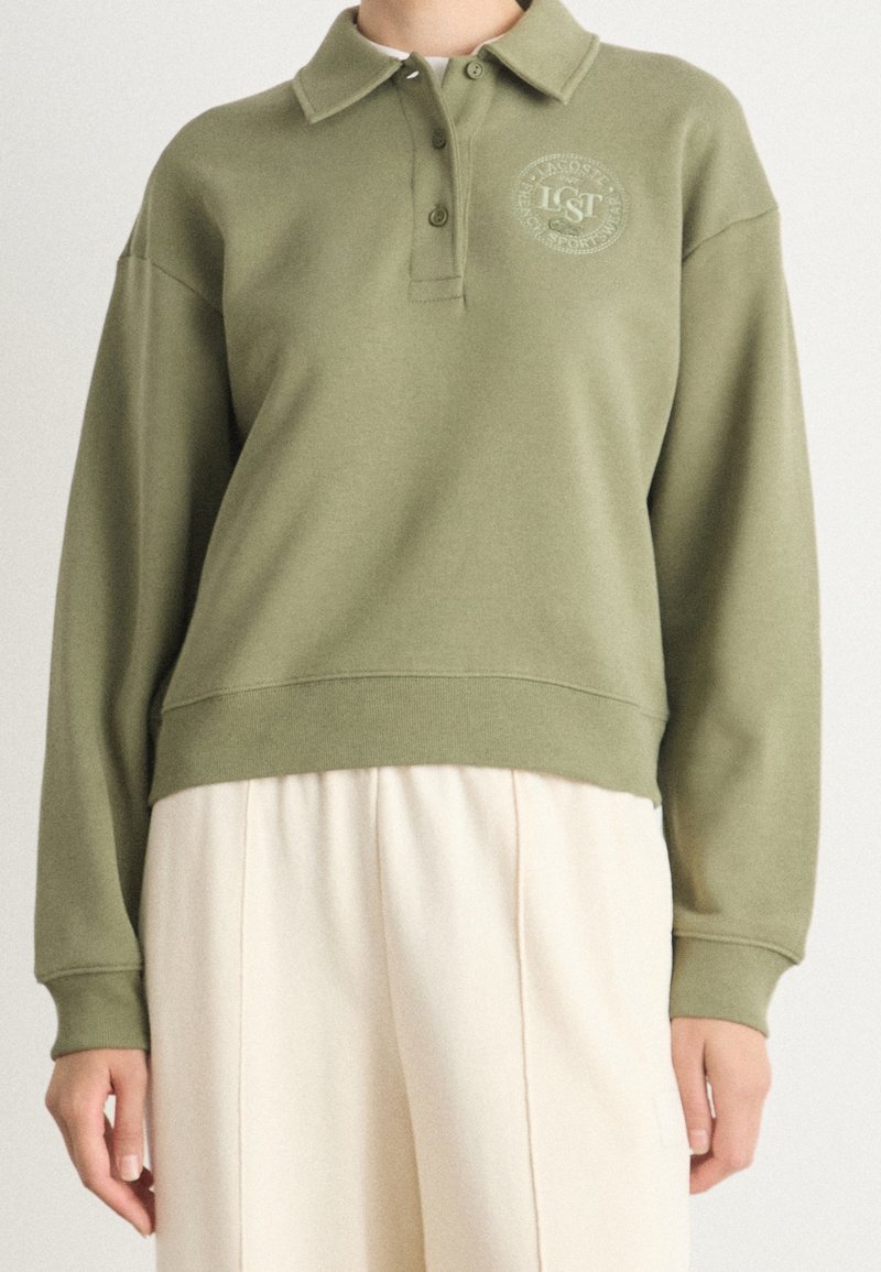 Personne portant un polo à manches longues vert olive avec un logo sur la poitrine et un pantalon ample de couleur blanc cassé, visible de l'épaule aux hanches.