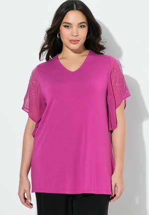 Frau mit dunklem Haar trägt ein leuchtend magentafarbenes Oberteil mit transparenten, verzierten kurzen Ärmeln vor weißem Hintergrund.