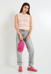Top sin mangas de punto con patrón de olas naranjas; jeans de cintura alta gris claro; gorra rosa brillante sostenida en la mano; zapatillas rosa.