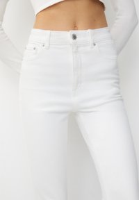 Detailní záběr na osobu, která má na sobě bílé džíny s vysokým pasem a bílé dlouhé crop tričko, které odhaluje břicho a obsahuje detaily knoflíků.