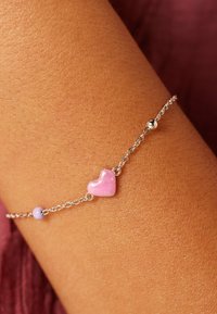Silver chain bracelet featuring a glossy pink heart charm and a small lavender bead, resting on a tanned wrist.