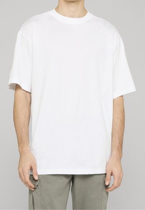 Person trägt ein schlichtes weißes T-Shirt mit kurzen Ärmeln und hellgraue Cargo-Hosen, steht vor einem neutralen grauen Hintergrund.