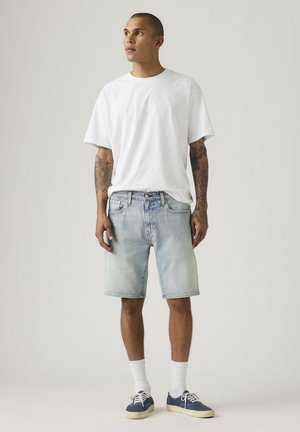 Hombre con tatuajes que lleva una camiseta blanca simple, pantalones cortos de mezclilla azul claro, calcetines blancos y zapatillas azul marino, de pie frente a un fondo neutro.