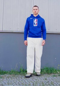 Niebieska bluza z kapturem z logo NBA, białe spodnie i szaro-zielone obuwie. Bluza ma kieszeń kangurką i ścianki z ribbingu. Jednolity tle.