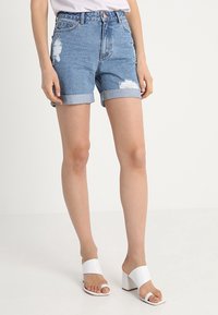 Džínové kraťasy světle modré barvy s opraným designem, s ohrnutými manžetami a předními kapsami. Nosené s bílými sandály na vysokém podpatku.