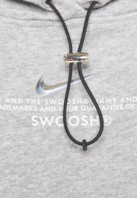 Grå hoodie med en borstad yta, som har en silverfärgad Nike-logotyp och svart snörning med en metallisk spärr. Vit text under logotypen.