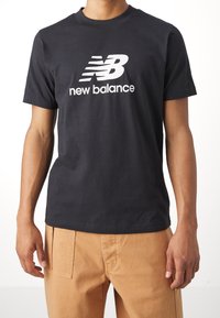 Camiseta de algodón negra con un gran logo blanco "NB" y el texto "new balance", cuello redondo y mangas cortas. Diseño simple, ajuste casual.