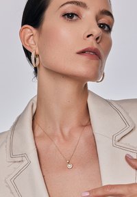 Złote kolczyki w kształcie kół zdobione klarownymi kryształami oraz delikatny złoty naszyjnik z okrągłym wisiorkiem na teksturowanym beżowym żakiecie.
