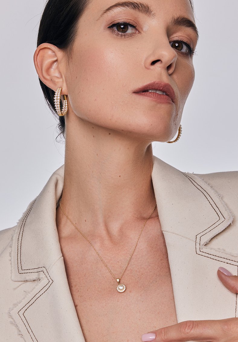 Złote kolczyki w kształcie kół zdobione klarownymi kryształami oraz delikatny złoty naszyjnik z okrągłym wisiorkiem na teksturowanym beżowym żakiecie.