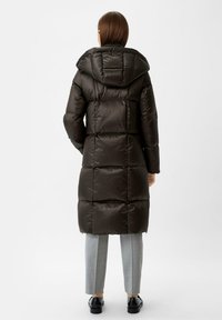 Lange pufferjas in donkerbruin, met een gewatteerd design en een capuchon, zachte textuur en elastische manchetten. Gecombineerd met grijze, op maat gemaakte broeken en schoenen.