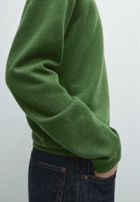 Zielony sweter z dzianiny z okrągłym dekoltem i prążkowanymi mankietami, zestawiony z ciemnymi dżinsami. Tekstura wygląda na miękką i ciepłą.