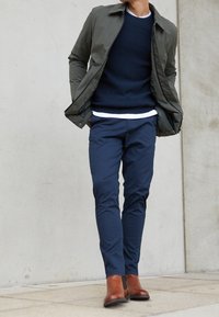 Olivgrüne Jacke, strukturiertes navy-blaues Sweatshirt, weißes Unterhemd, navy-blaue Hose, braune Leder-Stiefel bis zur Knöchel und vor einem hellen Betonschirm stehend.