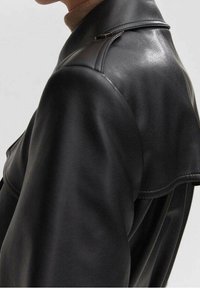 Schwarze Lederjacke mit glatter Textur, ausgestattet mit einem hohen Kragen und Schulterdetails. Das Design betont eine schlanke, enganliegende Silhouette.