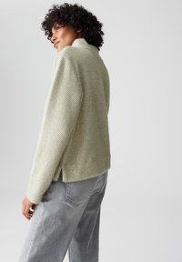 Gehaakte trui in lichtgroen met een textuurpatroon, voorzien van een hoge hals en zijsplitten. Gecombineerd met grijze jeans.