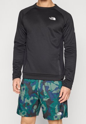Mann trägt ein schwarzes langärmeliges The North Face Shirt und grün-blau gemusterte Shorts mit The North Face-Logo auf beiden.