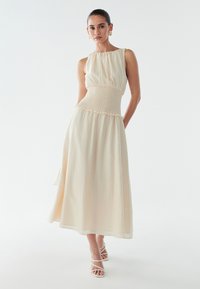 Robe beige clair avec un corsage smocké, sans manches, jupe fluide et ourlet à volants. Portée avec des talons blancs à lanières.