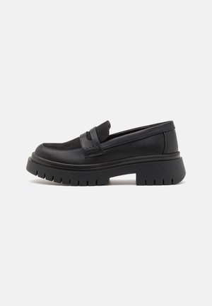 Mocassin en cuir noir avec une semelle épaisse crantée et un détail de bride sur le dessus, présenté de profil sur fond blanc.