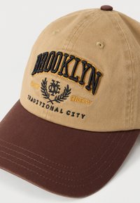 Khaki en bruine baseballpet met geborduurde letters "BROOKLYN", decoratieve elementen en contrasterende klep. Beige katoenen stof, gestructureerd ontwerp.