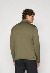 Chaqueta verde oliva con cremallera y alto cuello, textura suave y bolsillos laterales. Presenta un acento circular en el dobladillo. Pantalones negros llevados por debajo.