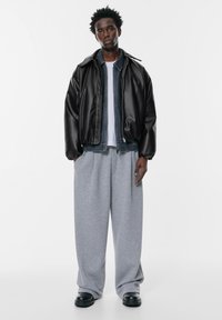 Črna usnjena jakna, sivi preveliki trenirki, bela majica, črne čevlje. Model stoji na enobarvni beli podlagi.