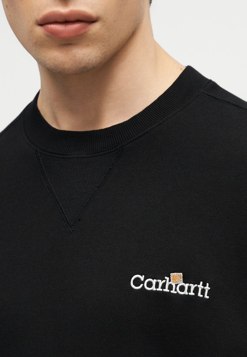 Primo piano di una persona che indossa una felpa nera Carhartt con logo ricamato bianco sul petto.