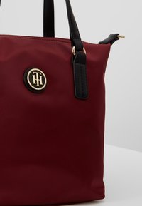 Borsa a mano rossa in nylon con manici neri, dettaglio logo dorato e forma strutturata. Presenta cuciture decorative e una texture liscia.