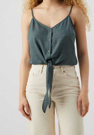 Kobieta ubrana w ciemnoszary bezrękawnik zapinany na guziki, zawiązany w węzeł w talii, w połączeniu z wysokim stanem kremowo-białych spodni.