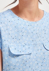 Blusa azzurra chiara in morbido tessuto, caratterizzata da un delicato motivo floreale, scollo rotondo e due tasche a pattina frontali.