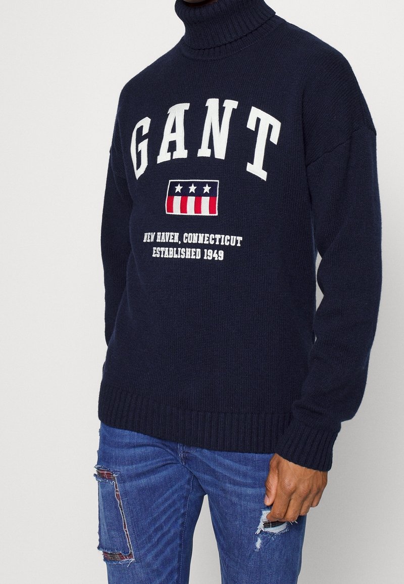 Navy Strick-Turtleneck-Pullover mit weißen "GANT"-Buchstaben und einem rot, weiß und blauen Logo. Rippbündchen und Saum, getragen mit verwaschenen blauen Jeans.
