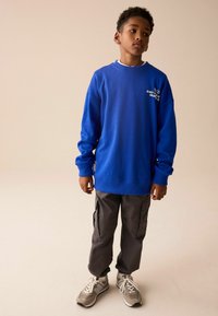 Blå sweatshirt med logotyp på bröstet, grå cargobyxor med sidofickor och grå sneakers med vita detaljer. Slät tygtextur.