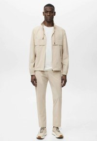 Beige letvægtsjakke med to brystlommer, parret med matchende beige bukser og hvid T-shirt. Skoene har neutrale toner og tekstur.