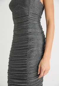 Robe grise en métal, ajustée, présentant un ruché vertical avec un tissu texturé. Design sans manches, silhouette moulante, coupe au niveau du genou.