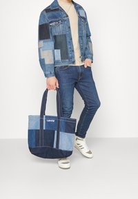 Denimová taška přes rameno se střihovým designem v různých odstínech modré, vybavená pevnými držadly a logem na přední straně.