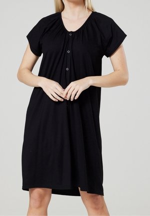 Woman wearing a loose black short-sleeve dress with a round neckline and three front buttons, standing against a plain background.