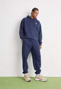 Mørkeblå bomullsblanding hettegenser med lommes foran og logo, parret med matchende joggebukser; fullført med beige sneakers og hvite sokker.