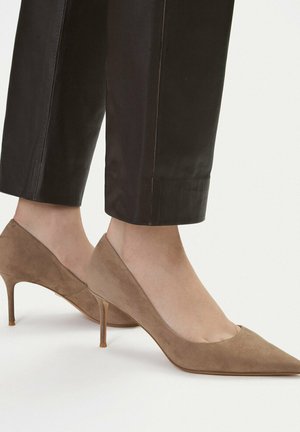 Piedi indossando décolleté a punta beige in camoscio abbinati a pantaloni neri in pelle tagliati sopra la caviglia su uno sfondo semplice.