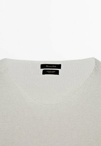 Pull en mélange coton-lin à col en V de couleur gris clair. Il présente un tricot texturé, avec des étiquettes de marque visibles sur le col, au design minimaliste.