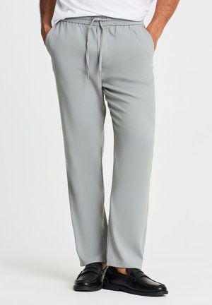 Uomo che indossa pantaloni grigio chiaro con coulisse, camicia bianca e scarpe nere senza lacci, in piedi con le mani nelle tasche su sfondo bianco.