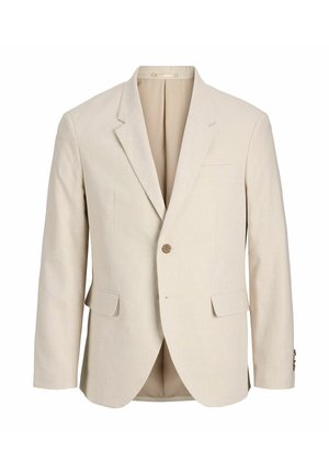 Unge person iført en beige dragt med hvid skjorte og hvide sneakers, stående og kiggende til siden mod en ensfarvet baggrund.