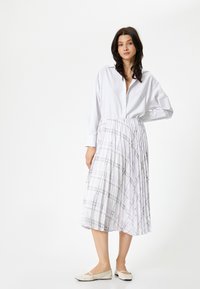 Chemise blanche à col classique associée à une jupe midi plissée arborant un léger motif à carreaux violets et des chaussures plates beiges.