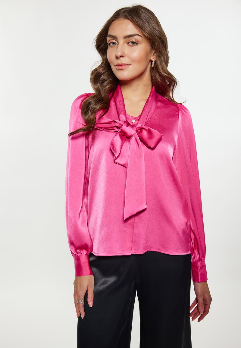 Blusa di raso di un vivace colore rosa con dettaglio a fiocco nella scollatura, maniche lunghe con chiusura a bottone e superficie liscia. Abbinata a pantaloni neri.
