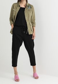 Oliivinvihreä napitettava takki taskuilla ja kukkaompeleilla, musta paita, mustat kapenevat housut ja vaaleanpunaiset slip-on-mule-kengät.