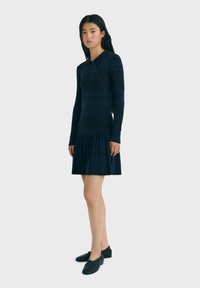 Neulemekko tummansinisenä, jossa on kauluksellinen kaula-aukko, pitkät hihat ja laskostettu hame. Ominaisuuksia ovat vaakasuorat raidat ja napit.