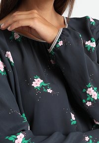 Blusa grigia scura con motivo floreale in rosa e verde. Presenta un colletto rotondo e polsini elastici, realizzata in tessuto leggero e strutturato.