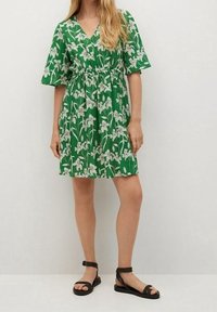 Grünes Blumenkleid mit V-Ausschnitt, kurzen schwingenden Ärmeln und elastischem Bund. Es hat ein weißes Blumenmuster und eine knielange Länge. Schwarze Sandalen.