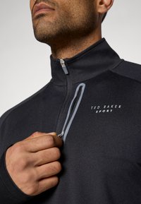 Jachetă sport neagră cu fermoar, cu guler înalt. Prezintă accente gri și logo-ul "TED BAKER SPORT" pe piept. Material neted, texturat.